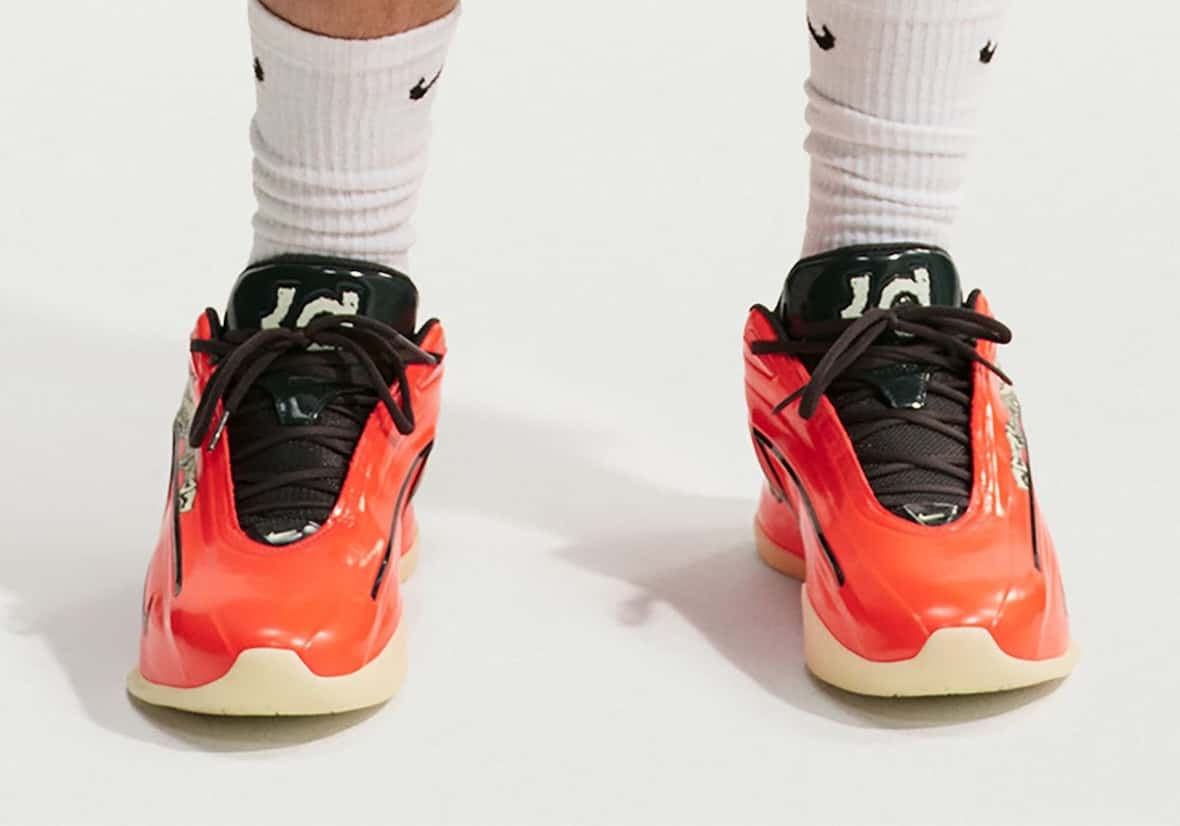 Vista frontal de un par de Nike KD 19 Halloween puestos con medias blancas Nike, con capellada naranja de acabado brillante, cordones marrón oscuro, lengüeta verde oscura con el logo KD en tono claro, un gráfico claro en el lateral y suela beige semitranslúcida, sobre fondo blanco